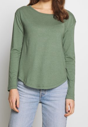 Femme portant un t-shirt à manches longues vert olive à col rond, associé à un jean bleu clair, debout devant un fond clair uni.