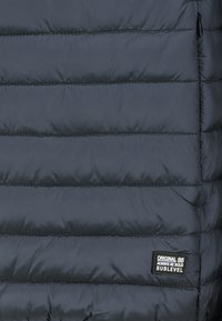 Marineblaue Steppjacke mit horizontalen Steppnähten und glatter Oberfläche. Verfügt über ein schwarzes rechteckiges Logotag mit weißem Text.