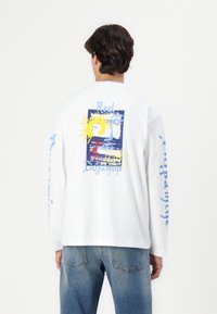 Tricou alb din bumbac cu mânecă lungă, prezentând un grafic colorat pe spate cu soare și designuri abstracte, inclusiv text albastru.