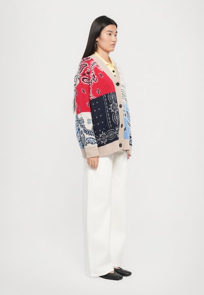 Gilet en tricot en patchwork de rouge, bleu, blanc et beige avec des motifs cachemire, doté d'une fermeture à boutons sur le devant et de manches larges. Associé à un pantalon blanc.
