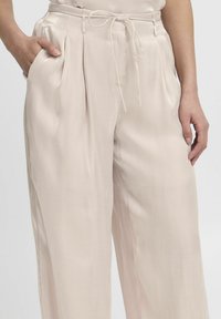 Wijdgevallen broeken van lichtbeige stof, met een trekkoord in de taille, zijzakken en plooien voor extra textuur en detail.