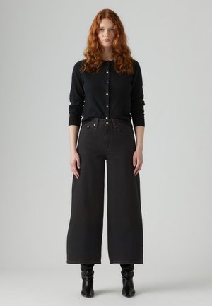 Black cardigan with round neckline and button closure, paired with high-waisted wide-leg black jeans. Model wears black ankle boots.