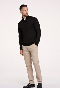 Maglione nero half-zip, design aderente, polsini a coste, indossato con pantaloni chinos beige. Scarpe nere lucide, texture liscia, sfondo neutro.