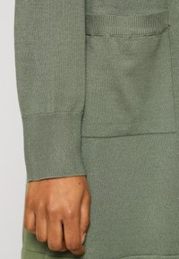 Handen vilar bredvid fickan på en ljus olivgrön stickad tröja med ribbad mudd och finstickad textur.