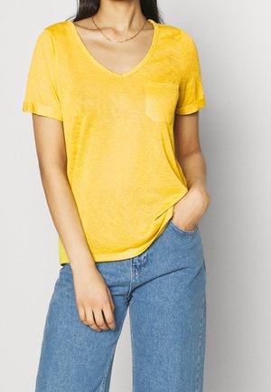 Frau trägt ein gelbes V-Ausschnitt-Kurzarm-T-Shirt mit Brusttasche und blaue Jeans, eine Hand in der Tasche und die andere entspannt.