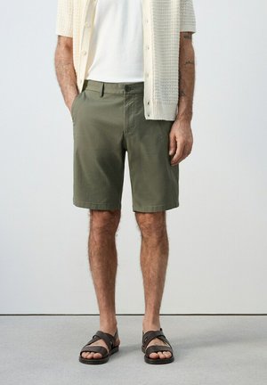 Hombre vistiendo pantalones cortos verde oliva, camiseta blanca, cárdigan de punto color crema y sandalias marrón oscuro, de pie con las manos en los bolsillos contra una pared lisa.