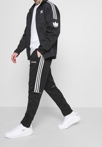 Svart träningsjacka med vita ränder, logotyp på bröstet; matchande svarta byxor med vita sidostripes. Vita sneakers full ändar outfiten.