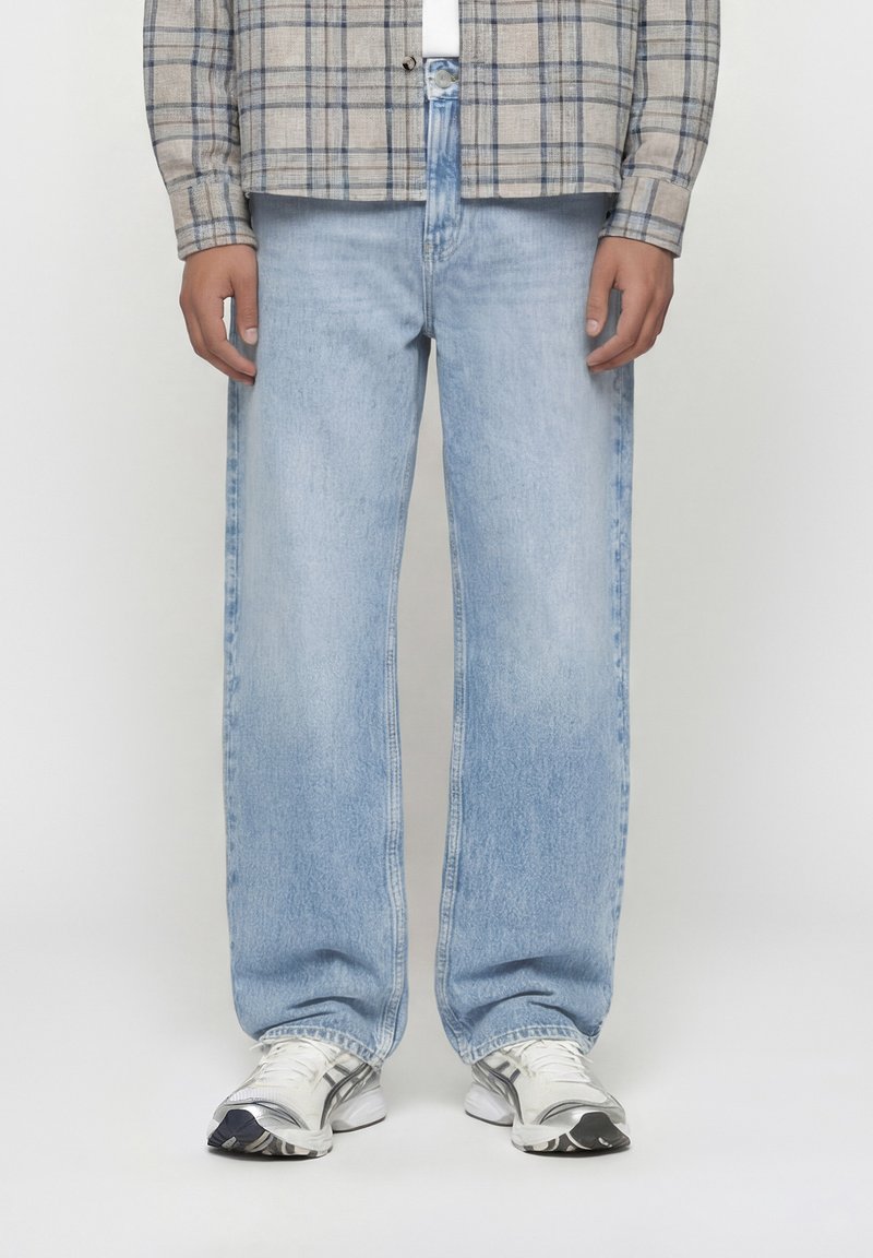 Άτομο που φοράει ανοιχτό μπλε τζιν παντελόνι φαρδύ, άσπρα και γκρι αθλητικά παπούτσια και μπεζ πουκάμισο καρό με κουμπιά, με τα χέρια χαλαρά στο πλάι.