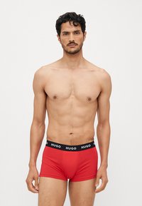 Boxers rouges en coton avec une ceinture noire arborant le logo "HUGO." La coupe est ajustée, mettant en valeur la silhouette du porteur.
