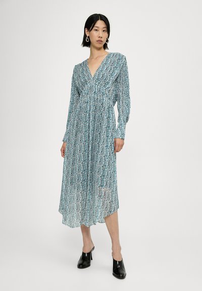 Vestido largo y transparente con un patrón de zigzag azul y blanco, con escote en V y mangas largas, combinado con mule de tacón alto negras.
