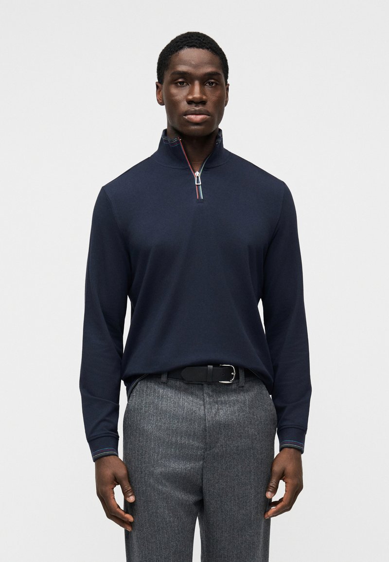 Μπλε σκούρο πόλο με μακριά μανίκια, φερμουάρ στο γιακά και πολύχρωμες ρίγες. Συνδυάζεται με γκρι υφασμάτινο παντελόνι και μαύρη ζώνη.