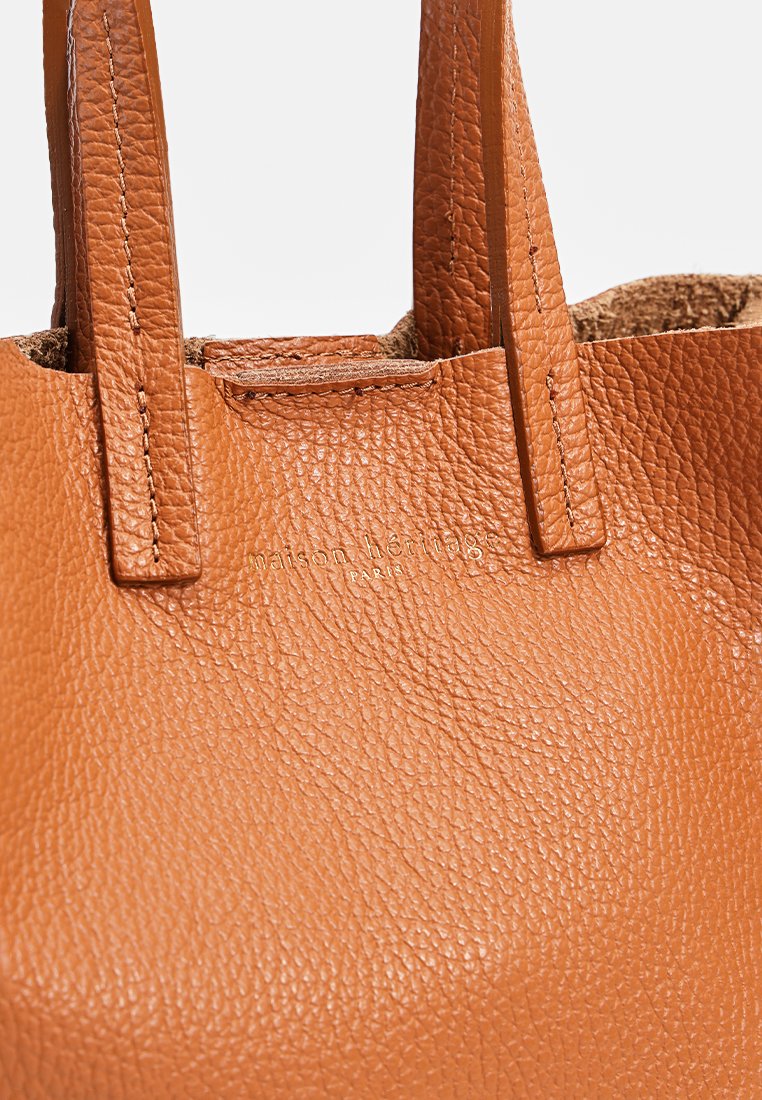 Tote-Bag aus braunem Leder mit strukturierter Oberfläche, ausgestattet mit kurzen Griffen und einem dezenten, geprägten Logo auf der Vorderseite. Kompaktes und minimalistisches Design.