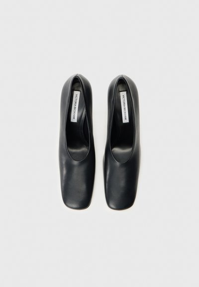 Čierne kožené topánky so štvorcovou špicou, plochým podpätkom a hladkým povrchom. Na vnútornej etikete je napísané "Victoria Beckham." Jednoduchý a minimalistický dizajn.