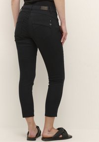 Jeans ajustados de mezclilla negra con cintura alta, que cuentan con bolsillos traseros, costuras sutiles y una textura suave. Llevados con sandalias negras.