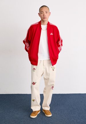 Rød idrettsjakke med hvite striper, hvit T-skjorte, kremfargede bukser dekorert med fargerike broderte mønstre, og brune joggesko.