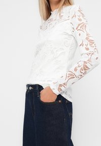 Bílý top s dlouhým rukávem s květinovým krajkovým vzorem a vysokým límcem. Kombinován s tmavými džíny z denimu, zobrazující boční kapsu a přiléhavý střih.