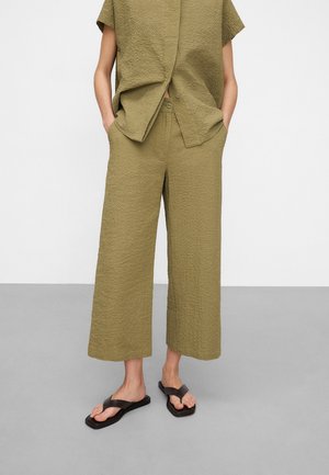 Personne portant un pantalon large court texturé vert olive et une chemise à manches courtes assortie, mains dans les poches, sandales noires à bout carré.