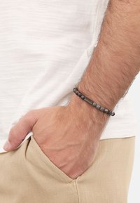 Bracciale di perle nere con pietre lisce e testurizzate, caratterizzato da accenti metallici. Indossato al polso, con la mano che riposa in tasca.
