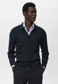 Cardigan a coste verde scuro con colletto, chiusura con bottoni e maniche lunghe, indossato sopra una camicia azzurro chiaro. Tessuto morbido e vestibilità su misura.