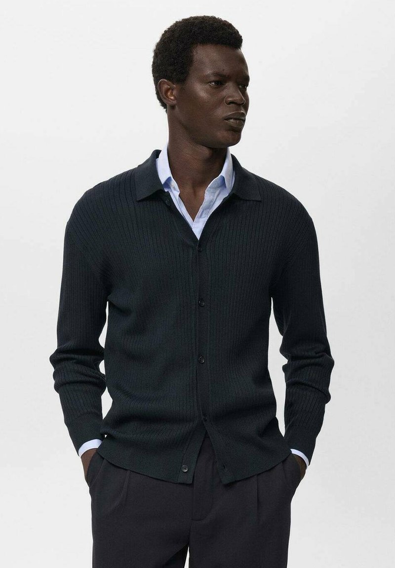 Cardigan a coste verde scuro con colletto, chiusura con bottoni e maniche lunghe, indossato sopra una camicia azzurro chiaro. Tessuto morbido e vestibilità su misura.