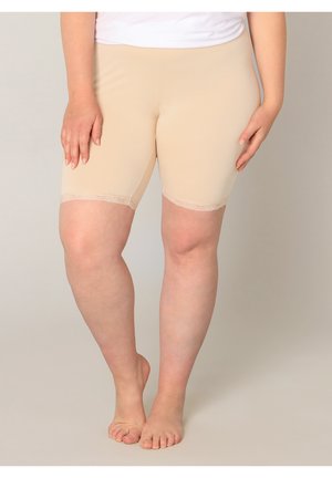 Persoon draagt beige knielange short met kanten rand, staat op blote voeten tegen een neutrale achtergrond.