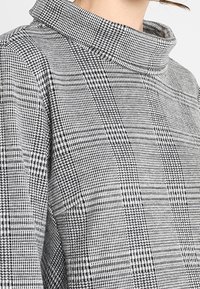  Suéter de cuello alto a rayas en gris y negro con tejido texturizado, que presenta un collar alto y un patrón de cuadrícula distintivo.