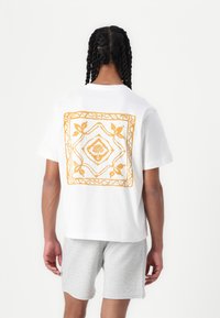 Camiseta blanca de manga corta con un diseño floral amarillo en la parte trasera, hecha de algodón suave y con un ajuste relajado.