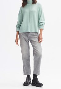 Vaaleanvihreä ylisuuri neulepaita korkealla kauluksella, ribbitetty pinta ja laskettu olkasauma, yhdistettynä harmaisiin suoriin farkkuihin ja mustiin nilkkasaappaisiin.