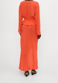 Langes, orange, fließendes Kleid aus leichtem Stoff, mit einem Peplum-Taille, langen Ärmeln und einem runden Ausschnitt. Flache, schwarze, geschlossene Sandalen mit Absatz.
