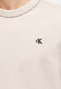 Κοντινό πλάνο μπεζ πουλόβερ με λαιμόκοψη παρεού και μαύρο κεντημένο λογότυπο "cK" στο αριστερό στήθος, με εμφανή ραφή στο γιακά.