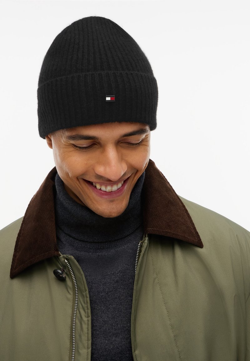 Homme portant un bonnet en laine noir, un pull à col roulé foncé et une veste verte avec un col marron, regardant vers le bas et souriant.