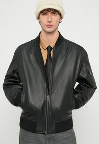 Melns ādas bomber jaka ar faktūras apdari, ribotu aproču un vidukļa joslu. Valkāta pāri brūnas krāsas kreklam un melnai kaklasaitei. Adīta krēmkrāsas cepure.