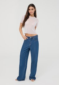 Top corto bianco con piccole brillantinature, abbinato a jeans a vita alta di colore blu e con gamba larga. La modella sta in piedi con una mano in tasca, su uno sfondo neutro.