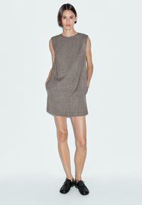 Sleeveless, brown shift dress with a relaxed fit, front pockets, and a subtle texture. Paired with black shoes. Smooth fabric finish.