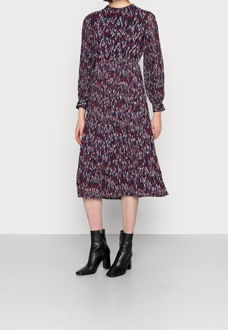 Femme vêtue d'une robe à manches longues et à motifs, longueur mi-mollet, associée à des bottines noires à talons, debout devant un fond uni.