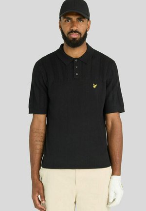 Mand med skæg iført sort kasket, sort strikket polo med korte ærmer og gul logo, beige bukser og hvid golfhandske på højre hånd.