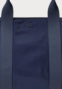 Sötétkék vászontáska tartós anyagból, két széles vállpánttal, a tetejénél középen egy kis gravírozott logóval. Simább tapintású.