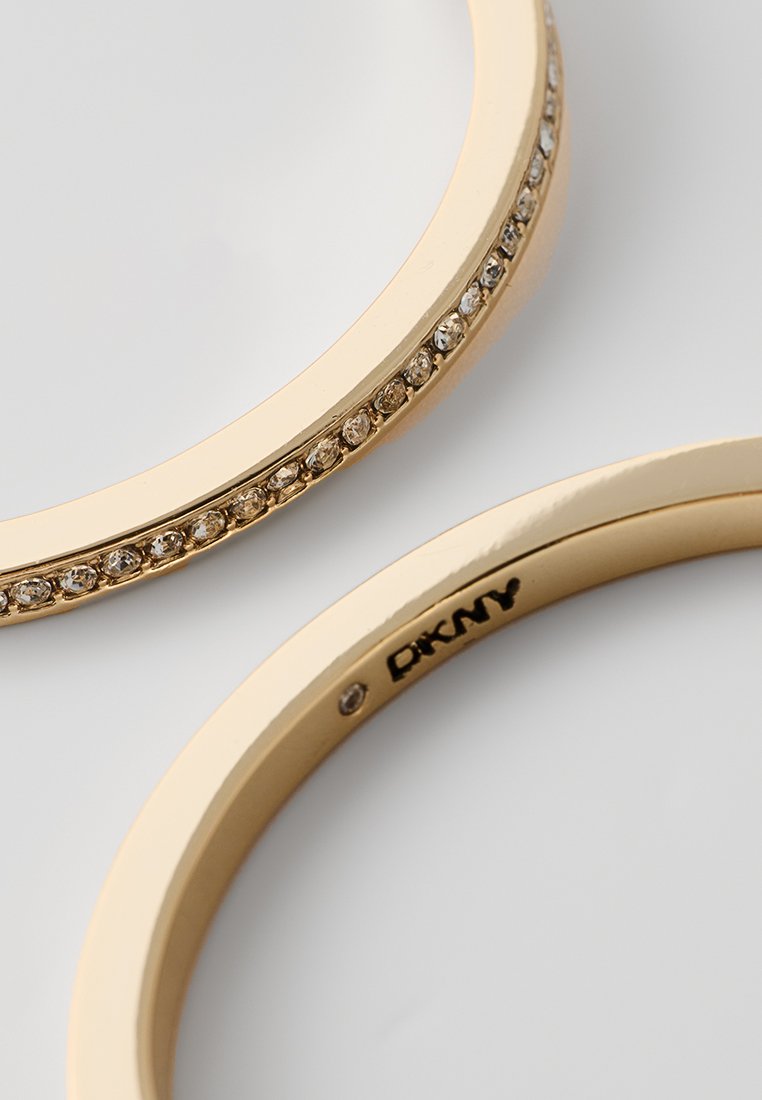 Brazaletes de color dorado con una superficie lisa y una fila de rhinestones a lo largo de un borde, con un logo de DKNY en el interior.