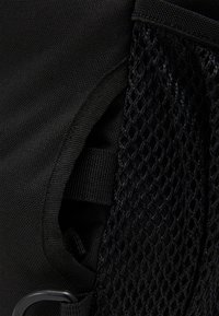 Sac à dos en tissu noir avec une pochette en mesh sur le côté, une sangle ajustable et une finition lisse. Couture durable visible le long des bords.
