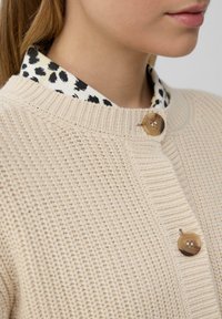 Giacche en tricot beige avec col côtelé et détails de boutons en bois. Superposé avec une chemise blanche présentant un motif à pois noirs.