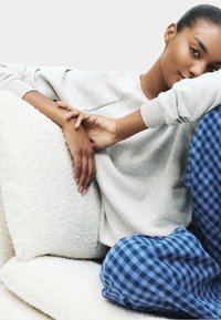 Junge Frau in grauem Sweatshirt und blauen karierten Hosen, die auf einer weißen strukturierten Couch liegt und ihren Kopf auf ihrem Arm ruht.