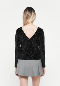 Schwarzes Samtoberteil mit langen Ärmeln, das ein V-Back-Design, ein strukturiertes florales Muster und eine figurbetonte Silhouette aufweist, kombiniert mit einem grauen Faltenrock.