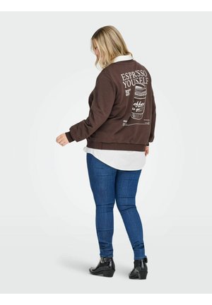 Brūna īsā džempera krekls ar tekstu "Izpaud savu espresso" un grafisku kafijas krūzi aizmugurē, virsū uz balta krekla, apvienots ar tumši ziliem džinsiem.