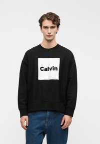 Mladý muž s kučeravými vlasmi, oblečený v čiernej mikine s nápisom "Calvin" v bielom štvorci, v kombinácii s modrými džínsami, stojaci pred jednoduchým pozadím.