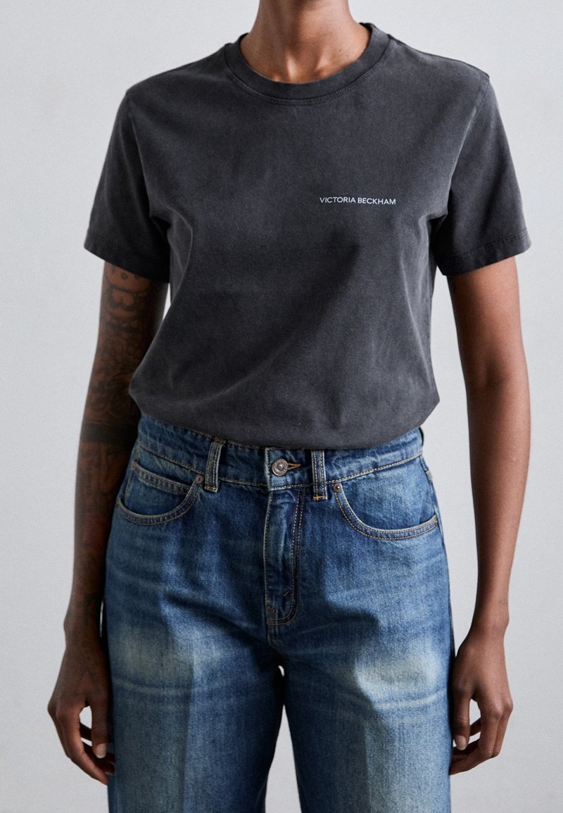 T-shirt nero in cotone a maniche corte con scollo rotondo, caratterizzato da un logo bianco "Victoria Beckham" sul petto, abbinato a jeans in denim blu.