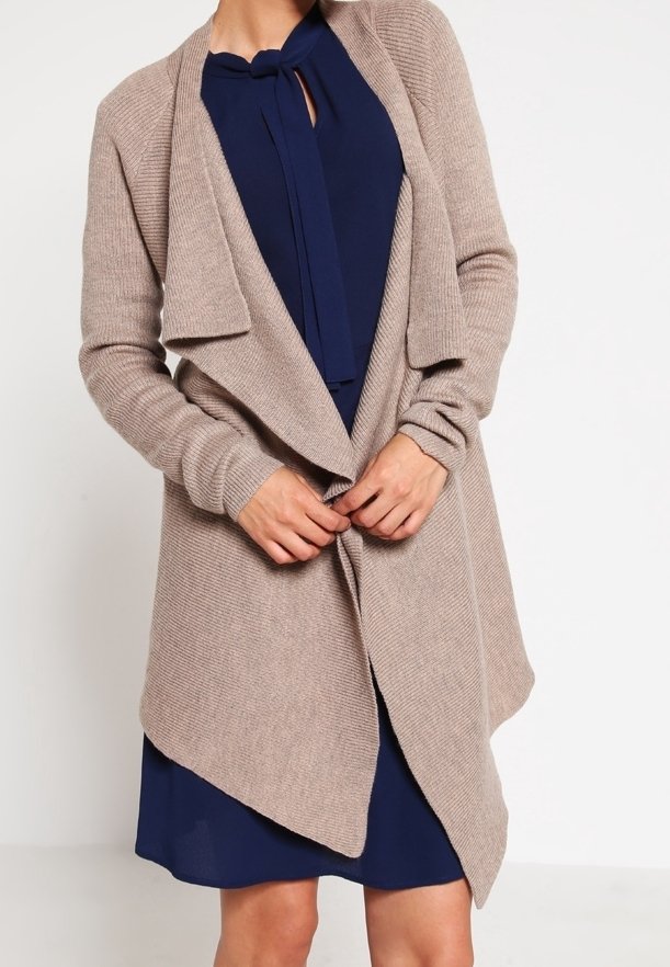 Cardigan lavorato a maglia beige con apertura frontale, design drappeggiato e texture a coste. Abbinato a una maglietta blu scuro con dettaglio a nodo sullo scollo.