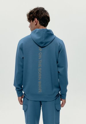 Muž otočený chrbtom, oblečený v modrej mikine s kapucňou a zodpovedajúcich nohaviciach, s vertikálnym nápisom "UP ON THE BORN WAVE" na chrbte mikiny.