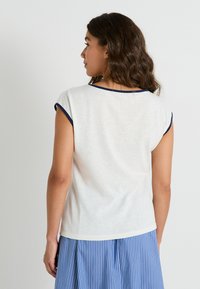 Top blanco de manga corta con ribete azul marino alrededor del escote y las mangas, que presenta un ajuste relajado. Textura suave; la vista trasera muestra un dobladillo redondeado.