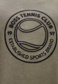 Vyšívaná nášivka s nápisom "BOSS TENNIS CLUB" a grafikou tenisovej loptičky. Farby: tmavomodrá na olivovozelenej textúrovanej látke.