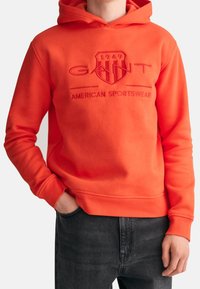 Helle orange Kapuzenjacke aus weichem Stoff, mit einem großen, gestickten Logo auf der Brust, gerippten Bündchen und einer Kapuze mit Kordelzug.